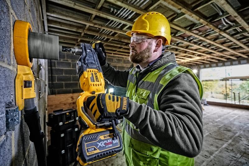 Ein Bauarbeiter mit gelbem Schutzhelm und Warnweste benutzt einen DEWALT DCD470N Akku-Bohrmaschine mit Lochsäge-Aufsatz, um in eine Betonwand auf einer Baustelle zu bohren.