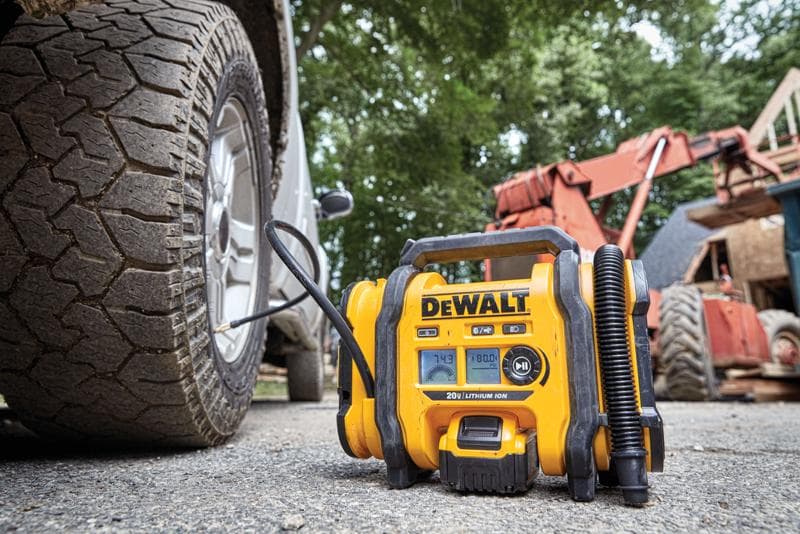 Ein DEWALT tragbarer Luftkompressor (Modell DCC020IB) wird im Freien verwendet, um einen Fahrzeugreifen aufzupumpen. Baumaschinen und Bäume sind im Hintergrund zu sehen.