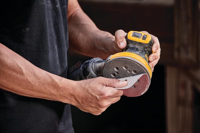 Eine Person hält einen kabellosen DEWALT Exzenterschleifer, Modell DCW210B, und befestigt ein Schleifpapier an dem Gerät.