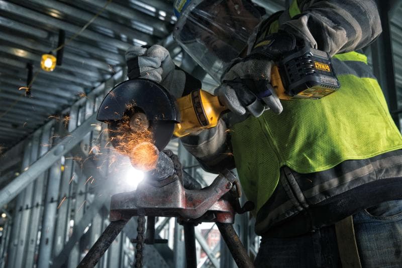 Nahaufnahme einer Person mit Schutzhandschuhen und Sicherheitsweste, die mit einem kabellosen DEWALT-Winkelschleifer Metall schneidet. Funken fliegen in einer industriellen Baustelle.