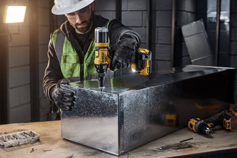 Eine Person mit Sicherheitsausrüstung verwendet ein kabelloses DEWALT DCF403NT Elektrowerkzeug, um an einer Metallbox in einer Industrie-Werkstatt zu arbeiten. Verschiedene Schrauben und Werkzeuge sind auf der Werkbank sichtbar.