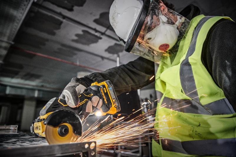 Eine Person mit Schutzausrüstung, darunter Helm, Gesichtsschutz, Maske, Handschuhe und Warnweste, verwendet ein kabelloses DEWALT DCS438N Elektrowerkzeug zum Schneiden von Metall, wodurch Funken entstehen.